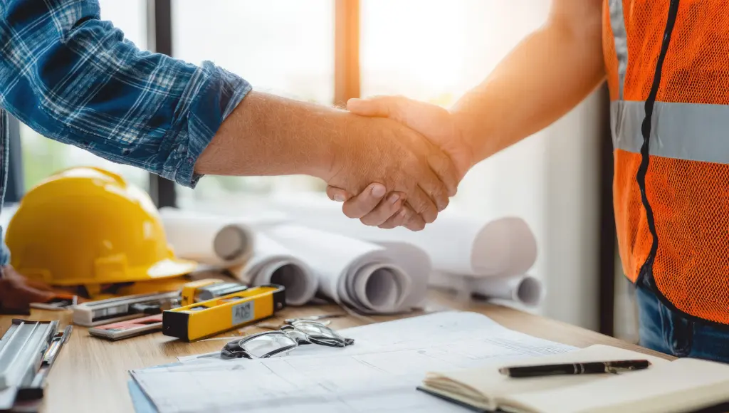 Two people shake hands over a table with blueprints, a yellow hard hat, and construction tools in view, suggesting an agreement on site.