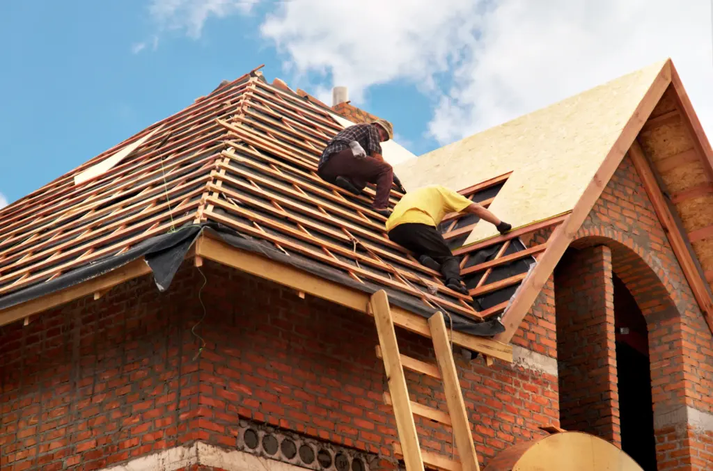 Construction workers on a steep wooden roof frame of a brick house, ladder leaning against wall, blue sky above.