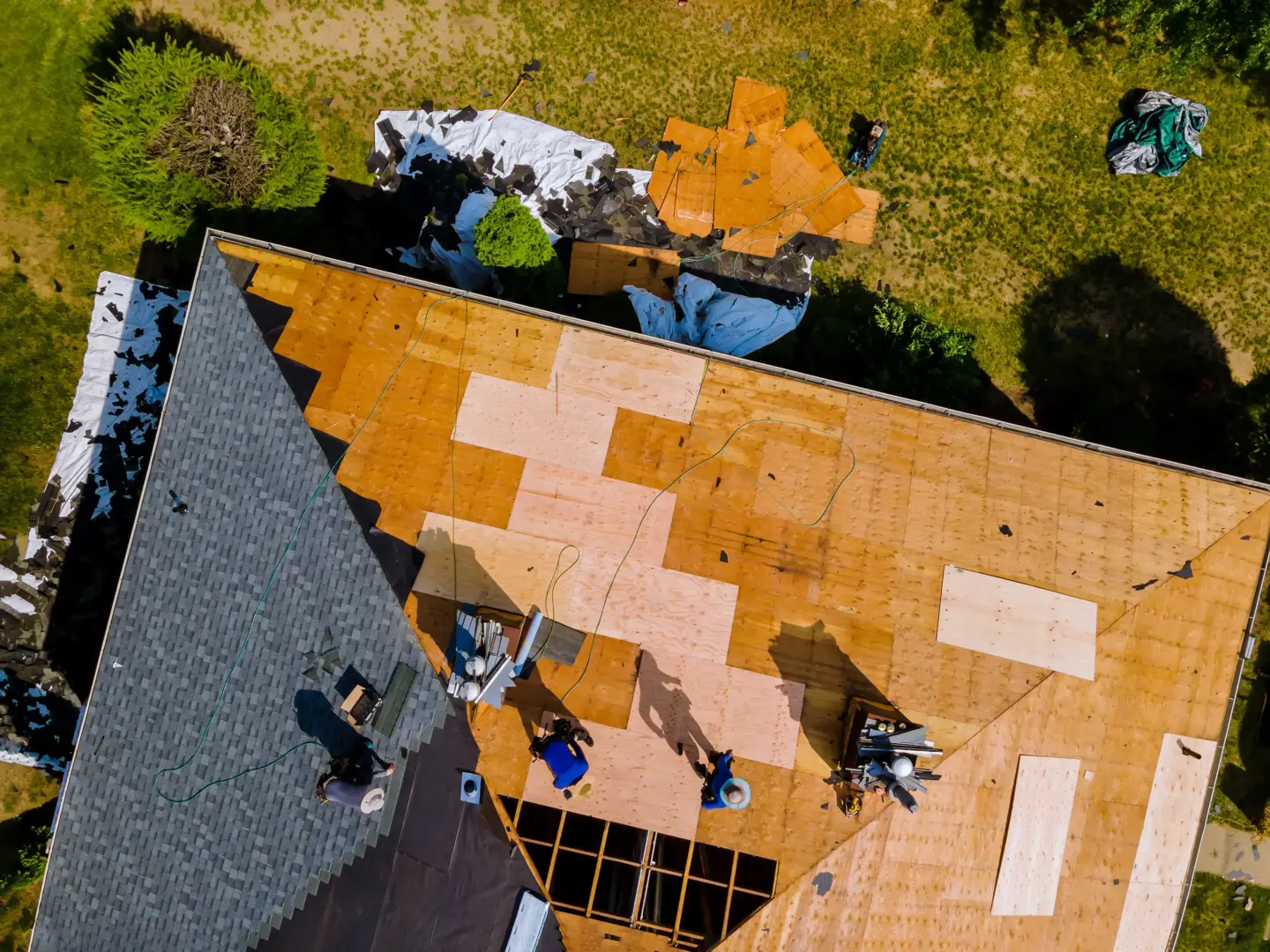 Aerial view of a roof under construction with wooden plywood sheets, tarps, and scattered tools; workers visible on the roof.