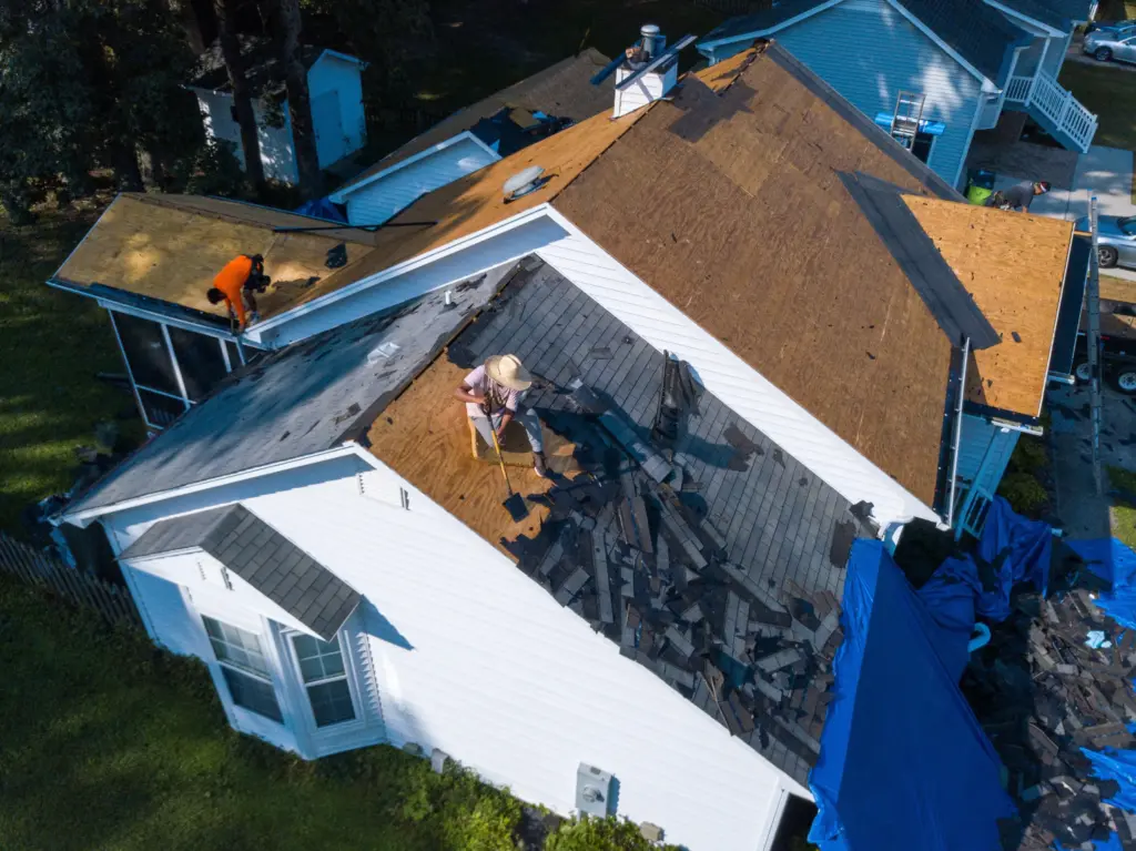 Aerial view of a house under roof renovation with workers on the sloped roof and scattered shingles and tarps nearby.