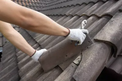 Person wearing white gloves uses a metal hook tool to grip brown roof tiles while climbing a sloped roof.