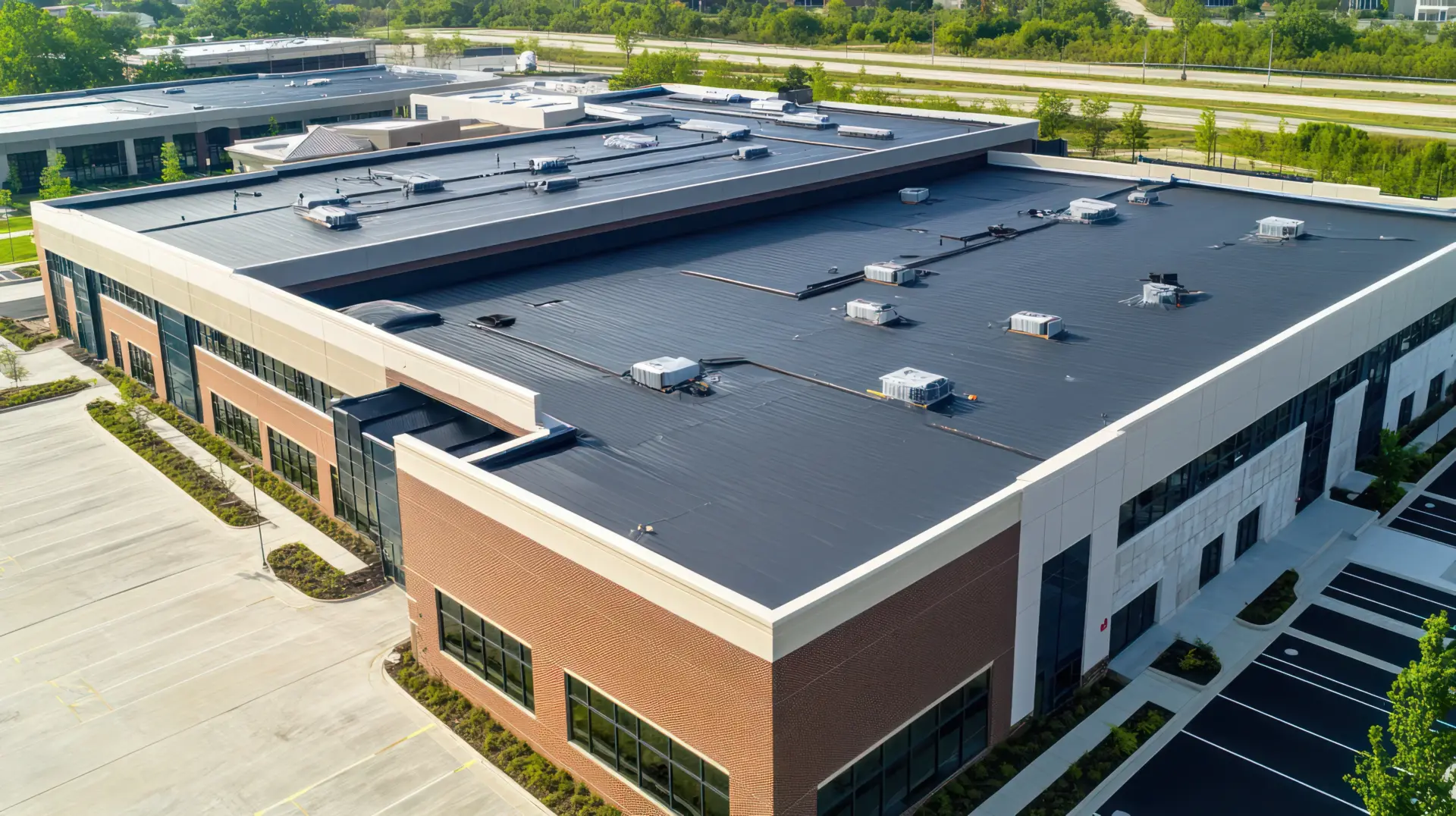 Aerial view of a modern commercial building with a large flat black roof containing HVAC units and vents, surrounded by greenery and parking.