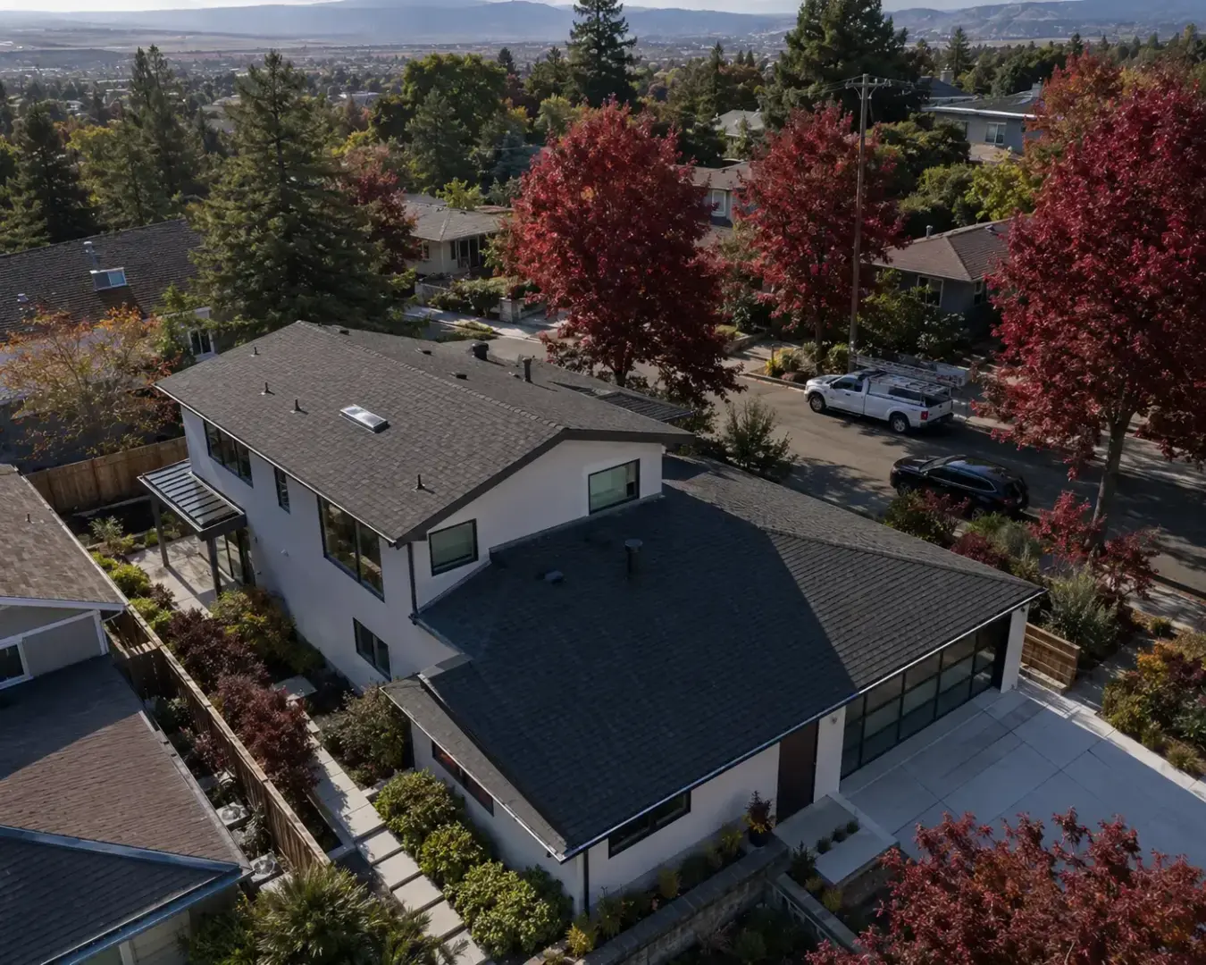 Aerial view of a modern white house with dark roof, surrounding trees, and a street with parked cars in a suburban neighborhood.