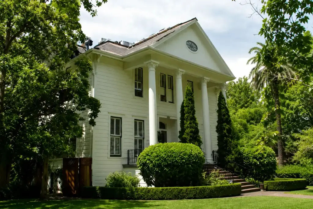 White two-story colonial house with a columned front porch, circular attic window, and lush landscaping around neatly trimmed hedges and trees.