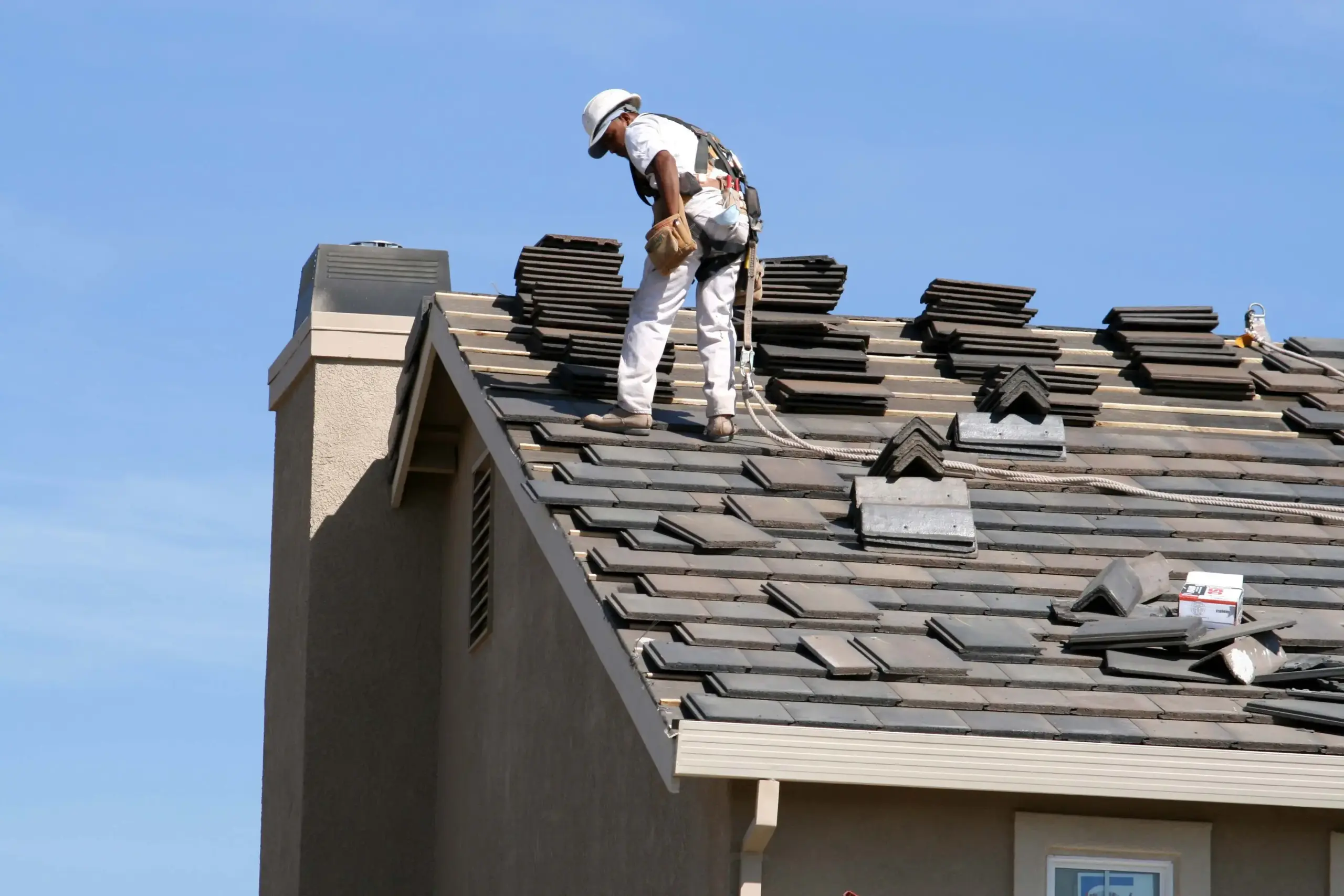 Construction worker in a white outfit and helmet on a sloped roof, harnessed with ropes and tools nearby