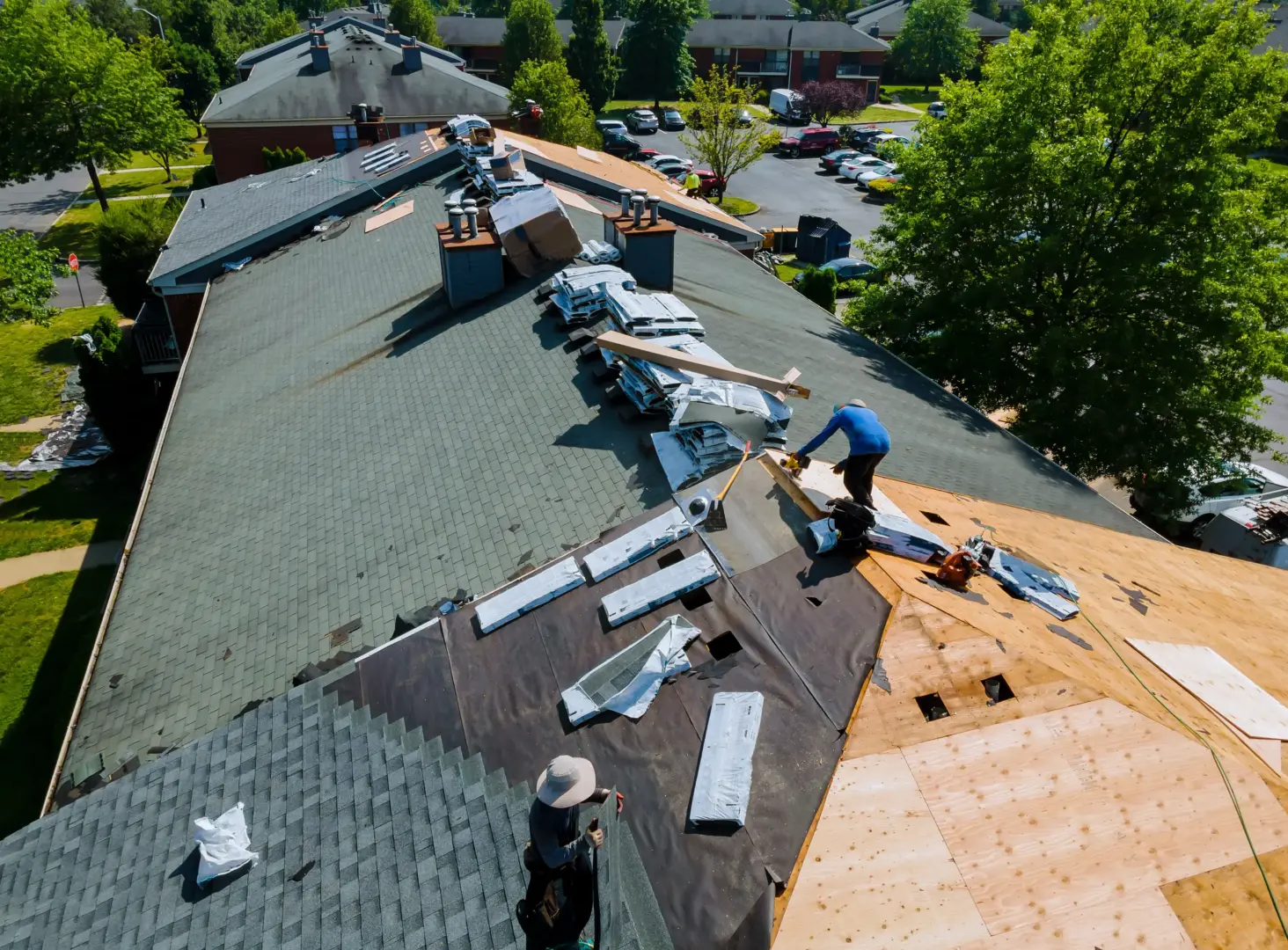 Aerial view of workers installing asphalt shingles on a large sloped roof with piles of materials nearby on a sunny day.