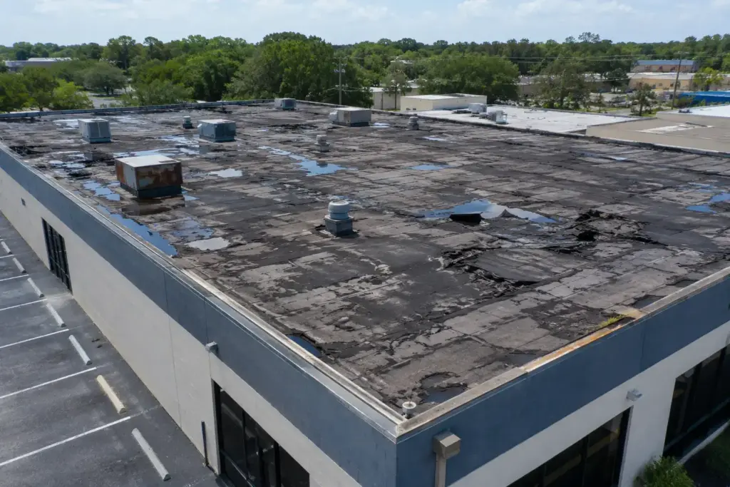 Flat commercial roof with cracked, peeling black membrane, several HVAC units, and scattered puddles of water.